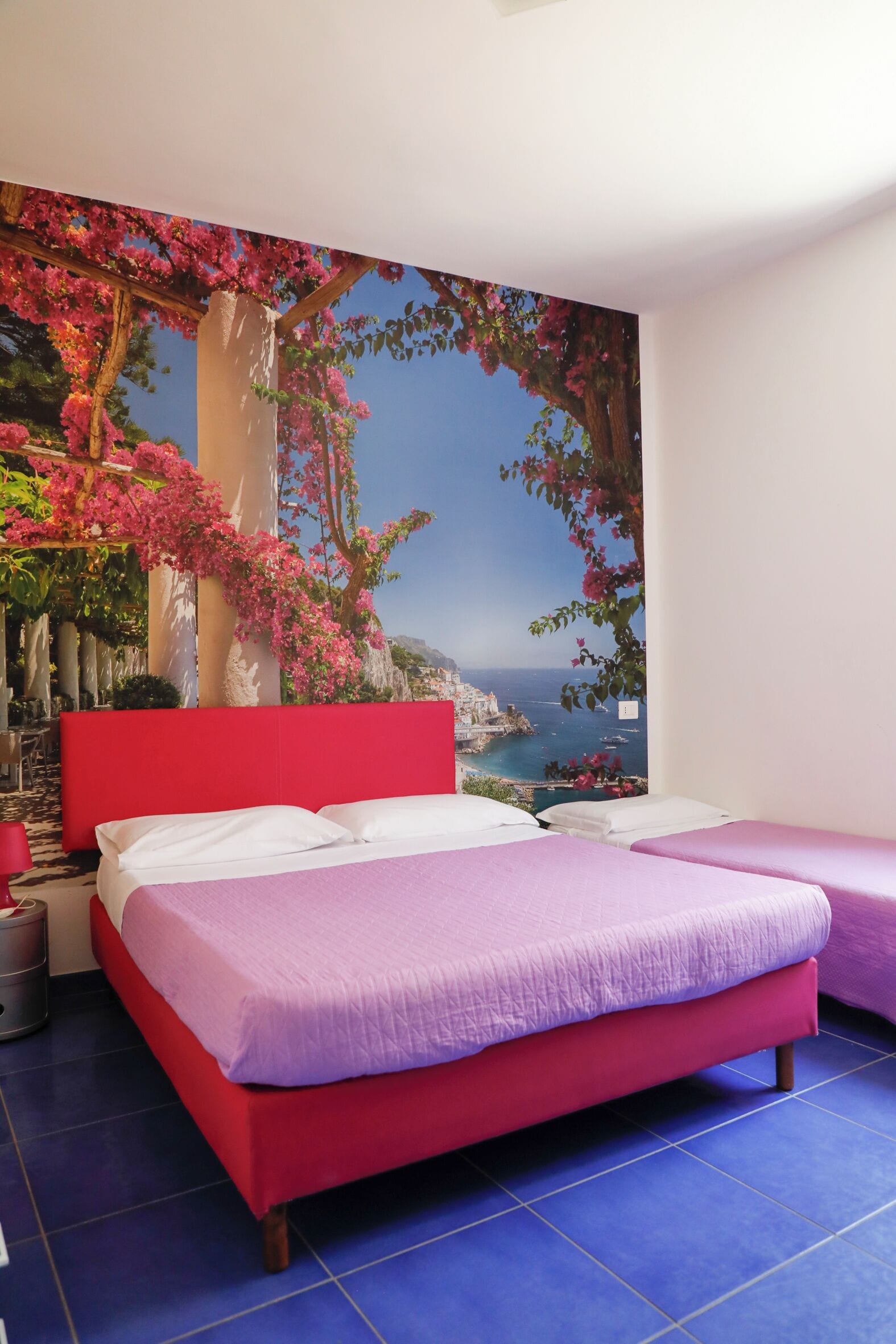 A bedroom with a red headboard, two beds with purple bedding, a large mural of a coastal landscape, and a blue tiled floor.