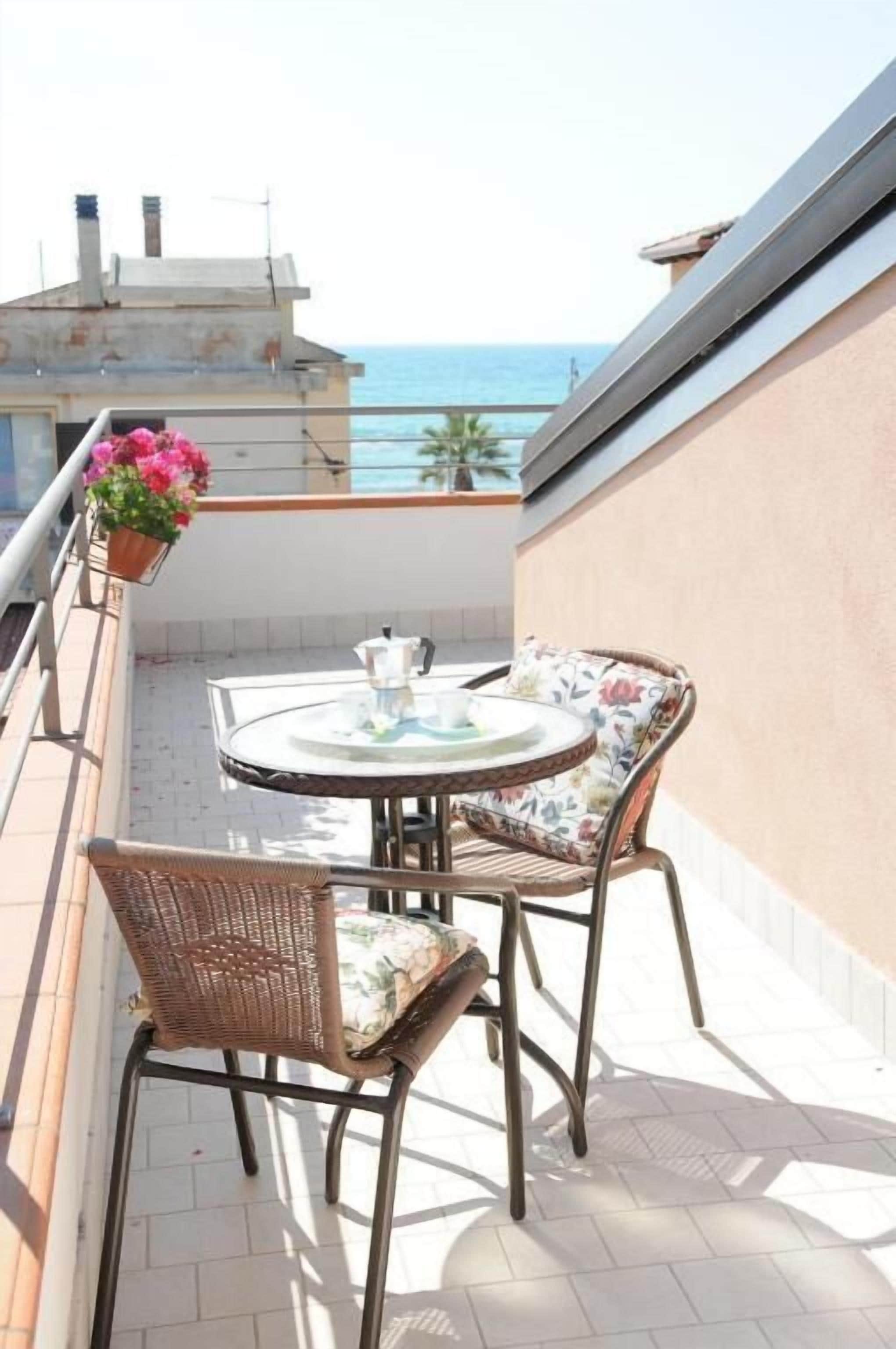 A small outdoor balcony with a round table and two wicker chairs, overlooking a body of water.