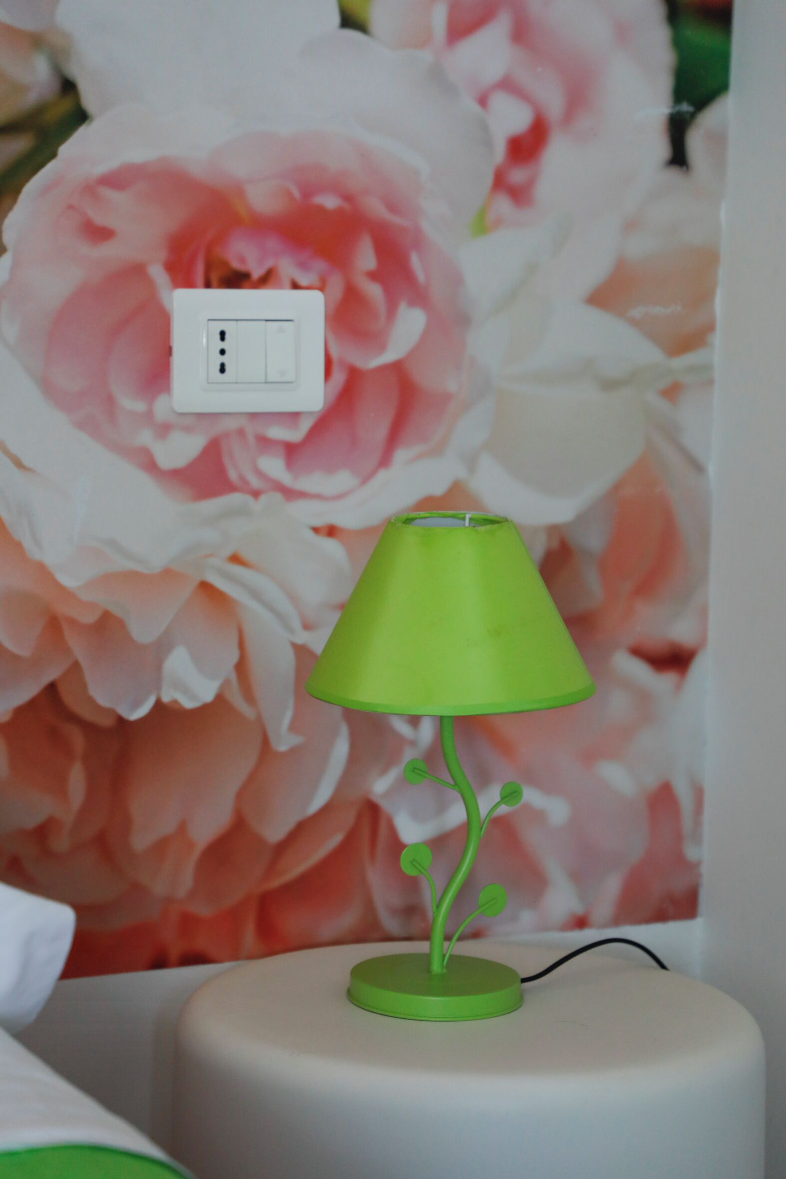 A green lamp with a plant-like base on a white table against a floral wallpaper background.