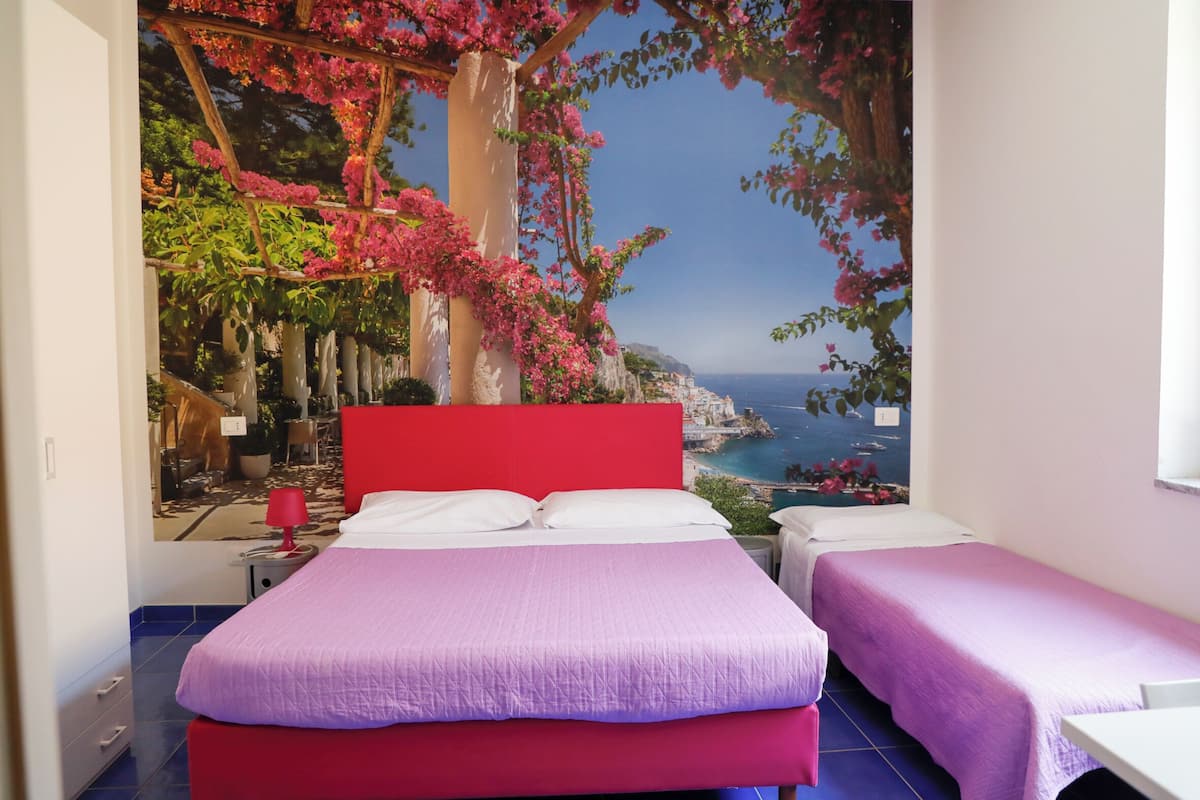 A bedroom with a large bed, two smaller beds, a red headboard, and a mural of a coastal scene.