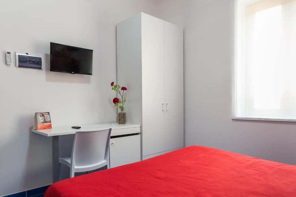 A bedroom with a bed, a desk with a chair, a TV, a wardrobe, and a window.