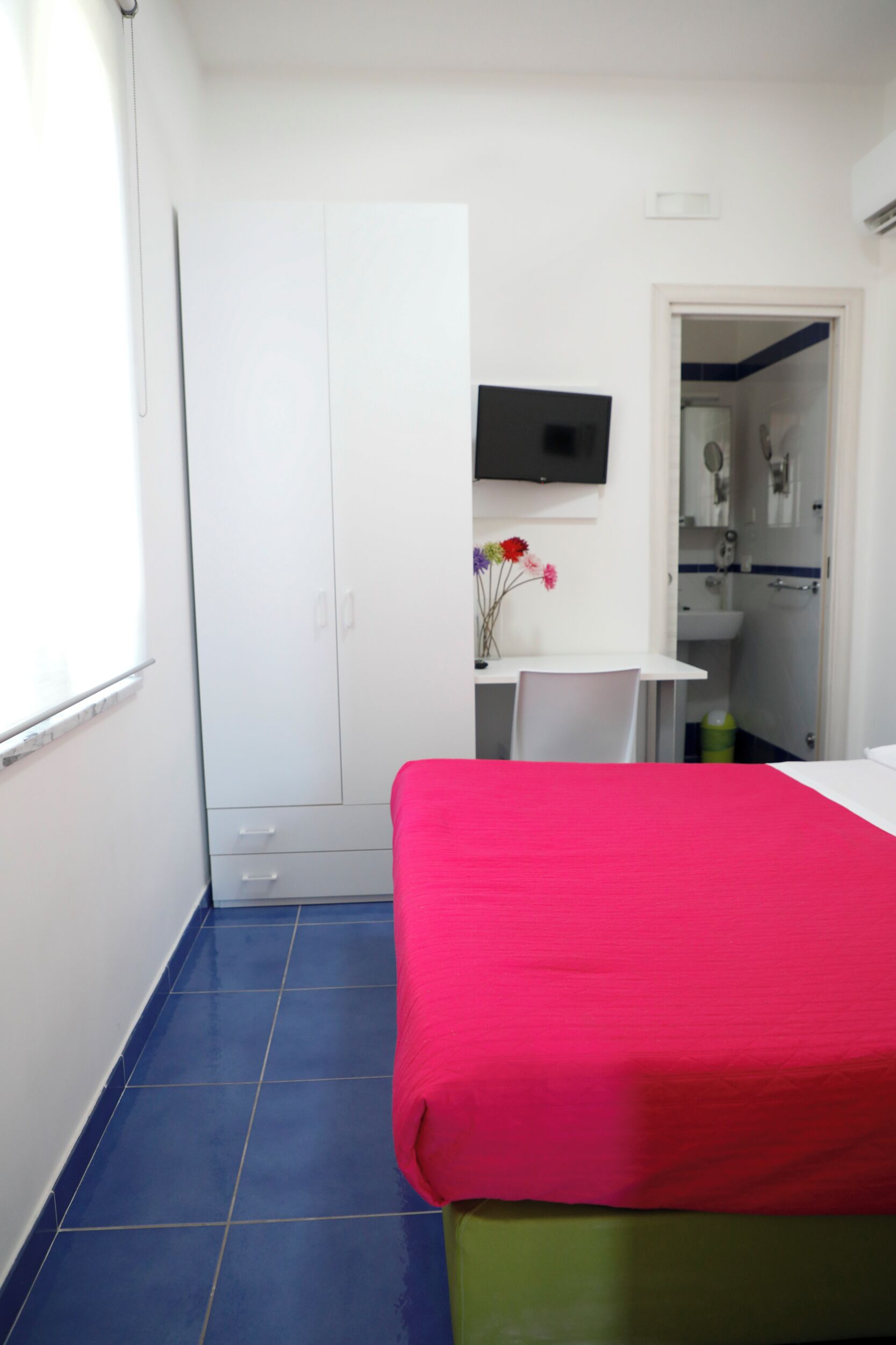 A bedroom with a bed, a desk with a chair, a TV, and a bathroom visible through an open door.