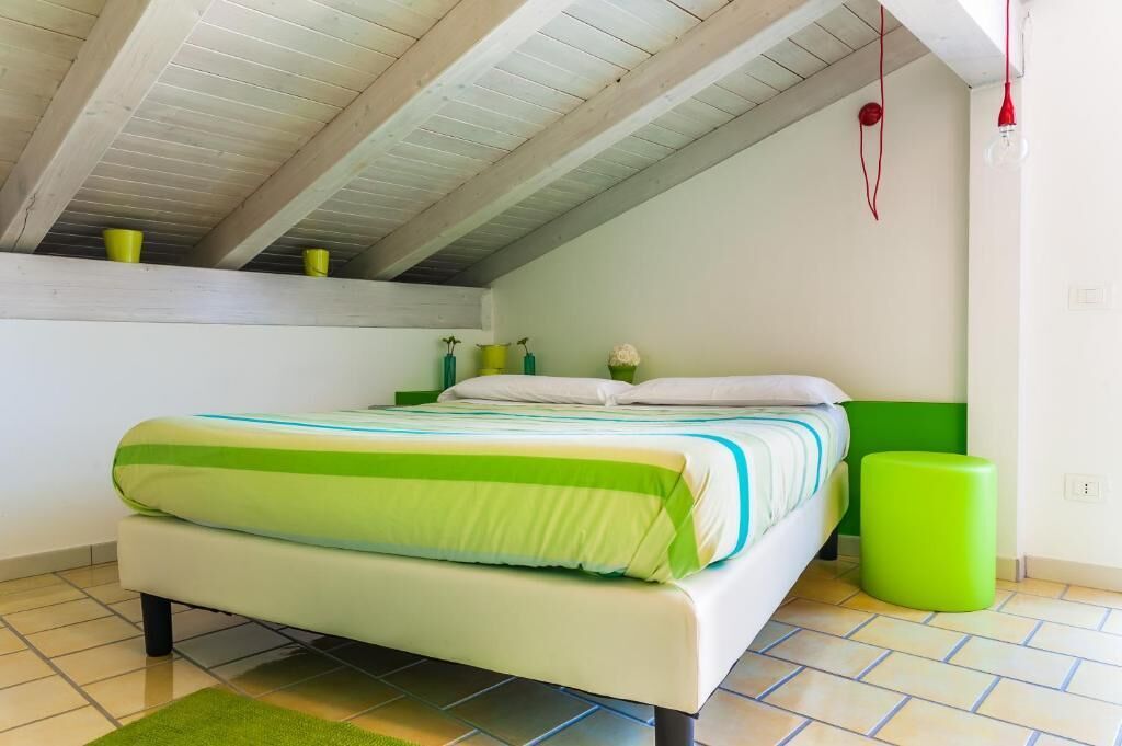 Un dormitorio con una cama, un taburete verde y una pequeña planta verde sobre una estantería.
