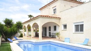 A swimming pool with a tiled edge, a patio with chairs, and a building with a tiled roof and arched windows. - Vacation home Dani by Interhome (L'Ampolla)