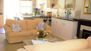 A living room with a beige sofa, a coffee table, and a fireplace. There is a kitchen in the background with a dining table and chairs. - Quaint family home close to Tram terminal (Franschhoek)