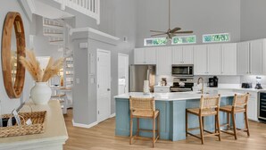 A spacious kitchen with white cabinets, a central island with bar stools, a ceiling fan, and a large window with green foliage. - Unique 3BR condo plus *LOFT* w/ golf cart & spacious open floor plan at Sandestin Golf & Beach Resort! (Miramar Beach)