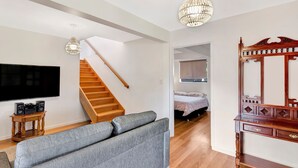 A modern living room with a grey sofa, wooden staircase, a TV, and a chandelier. - Hibiscus Dreaming - Brooms Head Village Location (Brooms Head)