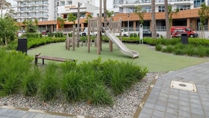 A playground with a slide and climbing structures, surrounded by greenery and a paved walkway.