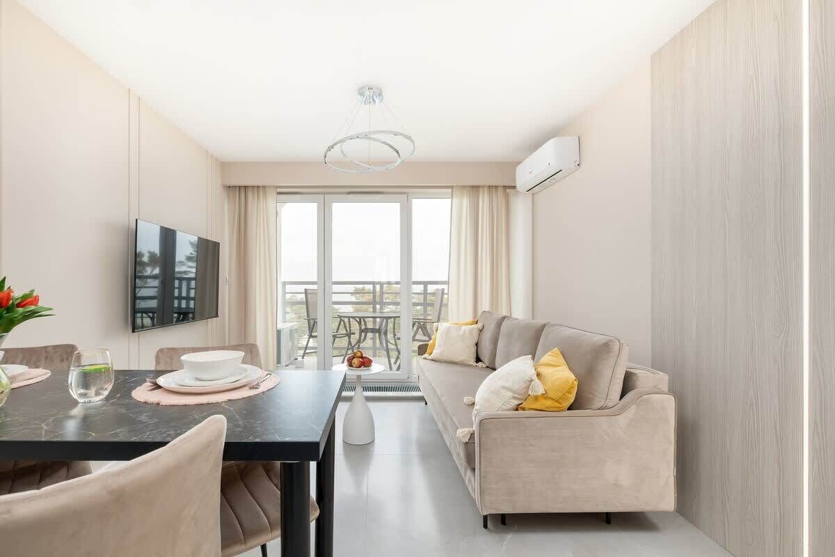 A modern living room with a sofa, a dining table, and a TV. There is a balcony with a view.
