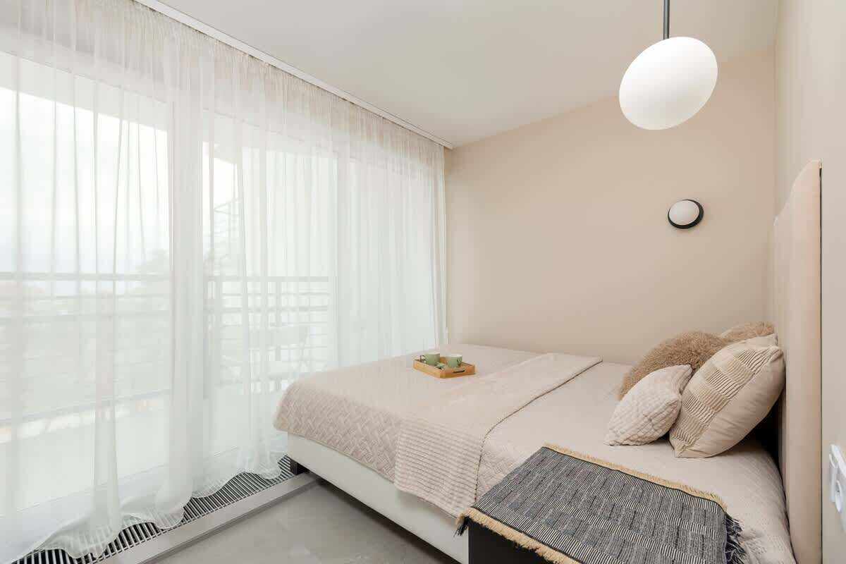 Quarto com cama, mesa de cabeceira, abajur e cortinas finas.