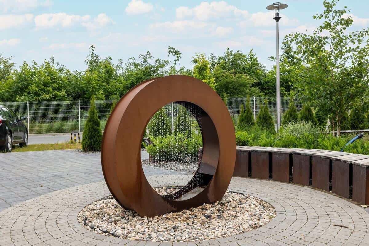 Una scultura metallica circolare con superficie riflettente, circondata da un'area pavimentata con ghiaia, con sullo sfondo vegetazione e una recinzione.