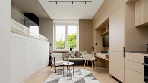 A modern kitchen with a built-in oven, a dining area with a sofa and chairs, and a large window providing a view of greenery outside. - Stylish Studio in the Heart of Warsaw | Close to Lazienki Krolewskie Park (Warszawa)