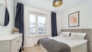 Un dormitorio con cama, cómoda, ventana con cortinas, radiador y un cuadro en la pared.