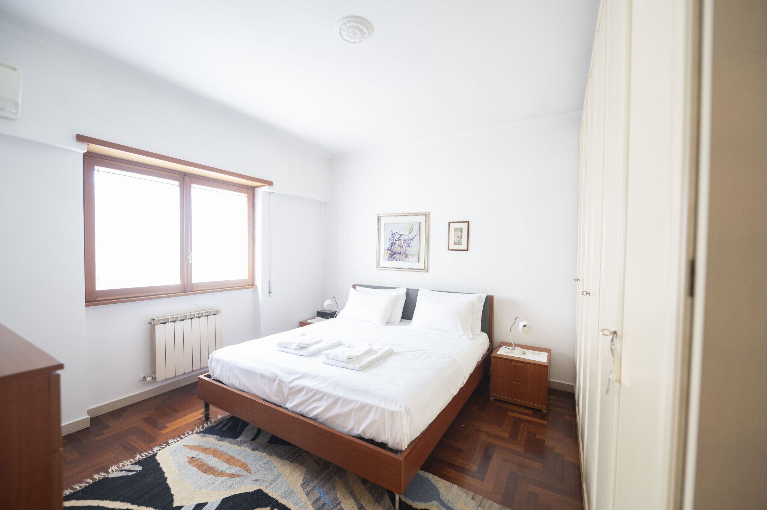 A bedroom with a wooden bed, a radiator, a window, a nightstand, and a bedside table.