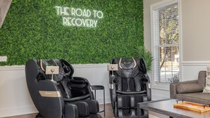 Een kamer met twee massagestoelen, een houten tafel met een dienblad, een bank en een muur met een groen wandbord waarop staat: THE ROAD TO RECOVERY.