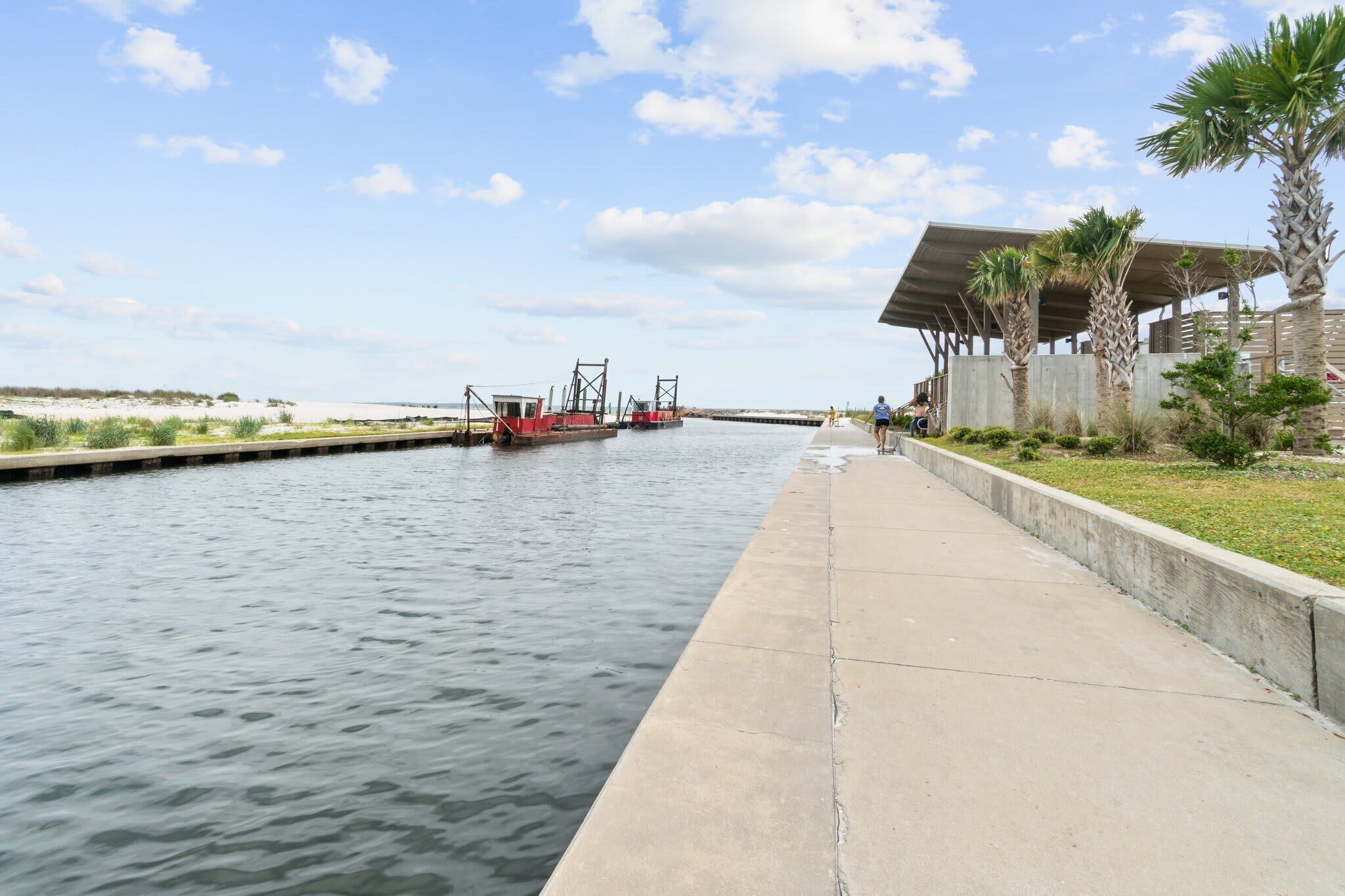 A canal with a boat dock, a concrete walkway, and a covered structure with palm trees.