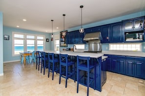 A kitchen with blue cabinets, a central island with bar stools, and a view of the ocean through large windows. - Sea Dragon - 6BR, Great Ocean Views, Pool, Hot Tub, Dog Friendly! (Surf City)