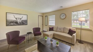A living room with a sectional sofa, two armchairs, a coffee table with plants, a wall clock, and a large framed picture of a bull. - Indoor fireplace with patio and pool table (Beaumont)