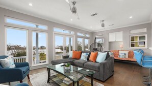 Ein geräumiges Wohnzimmer mit grauem Sofa, blauen Sesseln und einem Glastisch. Große Fenster bieten Ausblick ins Freie.
