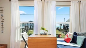 A room with a wooden table, a blue sofa, and a view of a house and beach through large windows with white curtains. - Welcome to Blue Jewel! (Galveston)