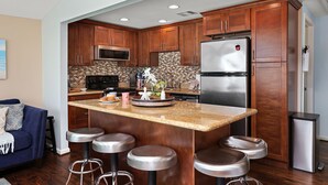A kitchen with wooden cabinets, a stainless steel refrigerator, a microwave, and a tiled backsplash. There are bar stools and a small island with a sink. - Welcome to Blue Jewel! (Galveston)