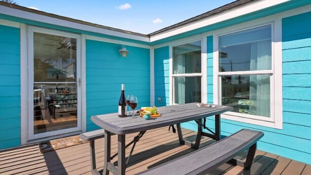 A turquoise house with a wooden deck, a table with a bottle of wine and fruit, and a bench. - Welcome to Blue Jewel! (Galveston)