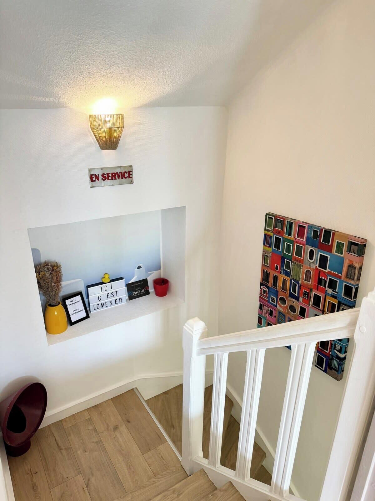 Een trap met een witte leuning, een wandlamp, een plank met decoratieve objecten en een kleurrijk kunstwerk.