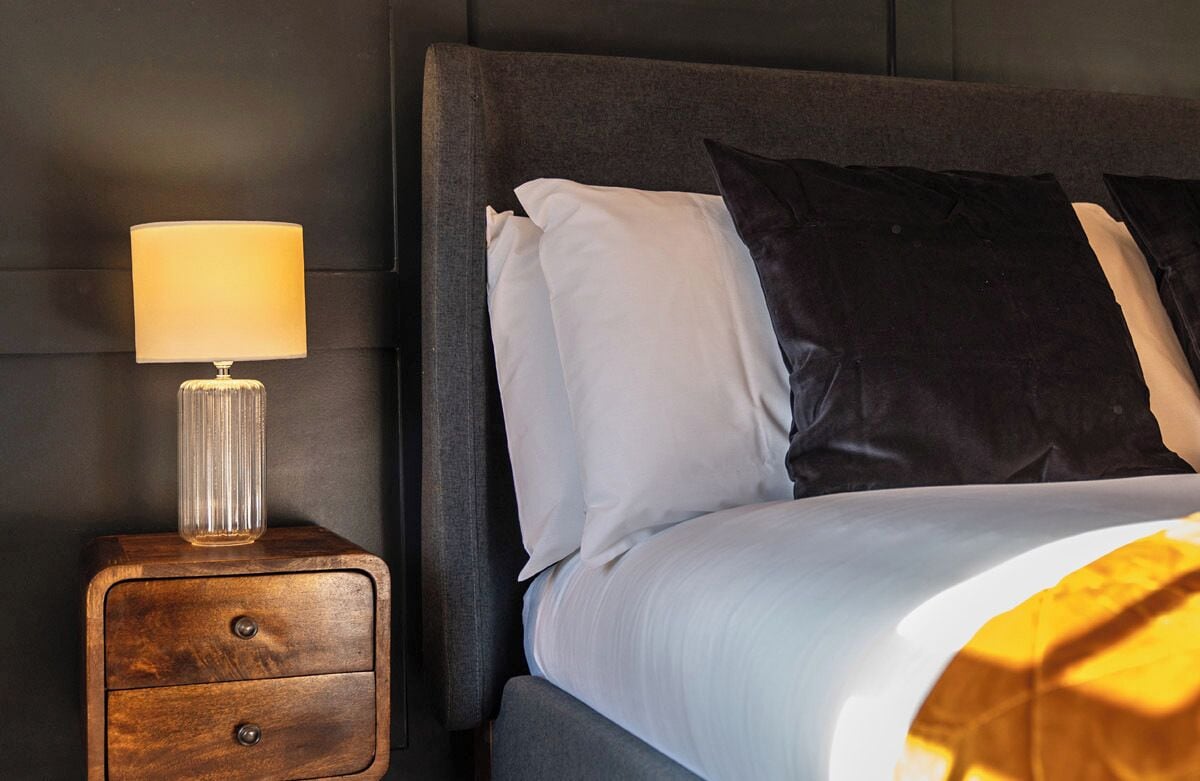 A neatly made bed with white and dark pillows, a wooden nightstand with a lamp, and a dark headboard.
