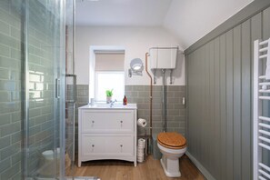 A bathroom with a glass shower enclosure, a white vanity with drawers, a toilet with a wooden seat, and a window with a view of greenery.