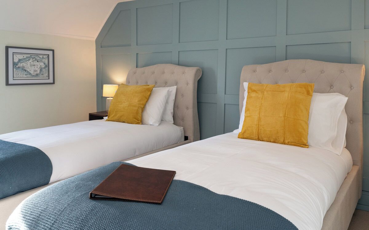 Une chambre d’hôtel avec deux lits, chacun doté d’une tête de lit, de draps blancs et d’un plaid bleu. On aperçoit un objet en cuir marron sur le lit, au premier plan.
