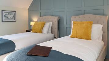 Une chambre d’hôtel avec deux lits, chacun doté d’une tête de lit, de draps blancs et d’un plaid bleu. On aperçoit un objet en cuir marron sur le lit, au premier plan.