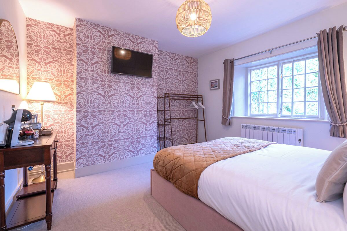 A bedroom with patterned wallpaper, a bed with a quilt, a TV mounted on the wall, a radiator, and a window with curtains.