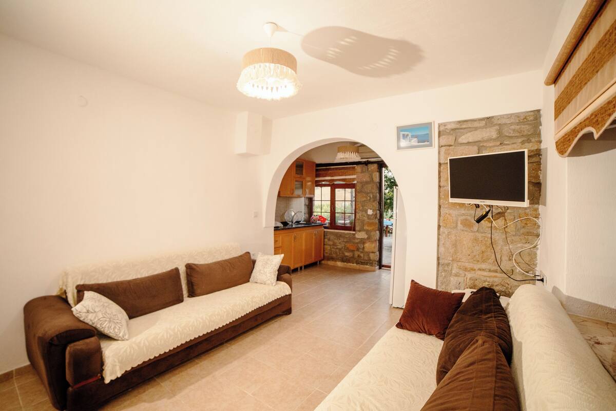 A living room with a sofa, a television mounted on a stone wall, and an arched doorway leading to a kitchen.