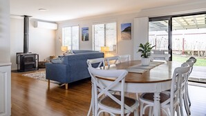 A cozy living room with a sofa, dining table, and chairs, a fireplace, and large windows with blinds. - Sandy Cottage in Narrawallee (Narrawallee)
