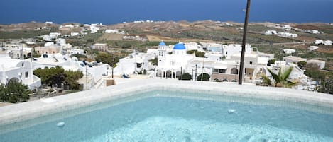 A swimming pool with clear blue water, surrounded by white buildings with blue domes, set against a backdrop of a coastal landscape.