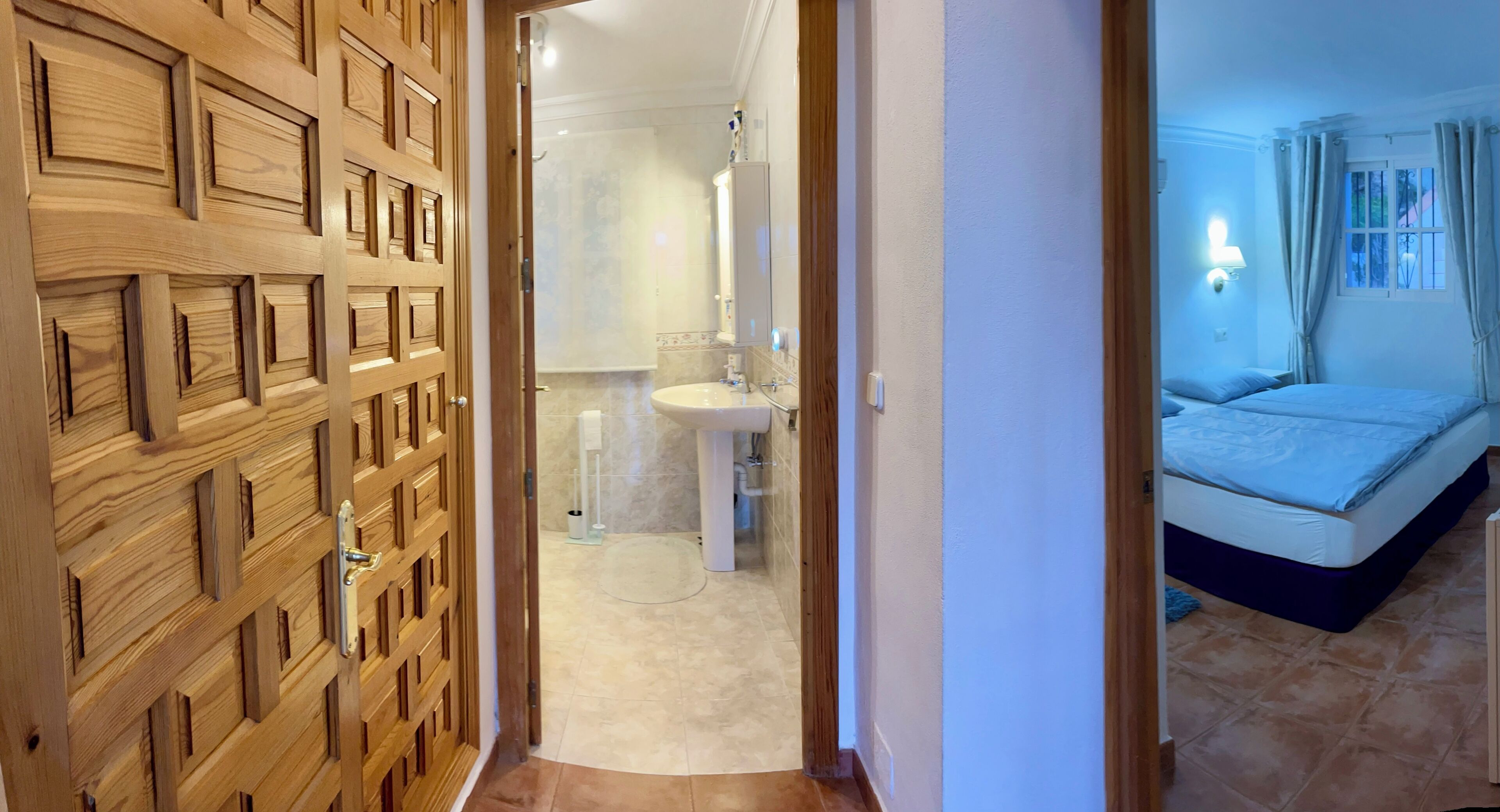 A hotel room with a wooden door, a bathroom with a sink and mirror, and a bed with a blue blanket.