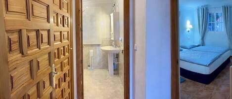 A hotel room with a wooden door, a bathroom with a sink and mirror, and a bed with a blue blanket.