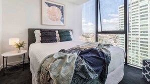 Una camera da letto moderna con un letto grande, un comodino con una lampada, un'opera d'arte incorniciata a parete e vista sulla città attraverso ampie finestre.