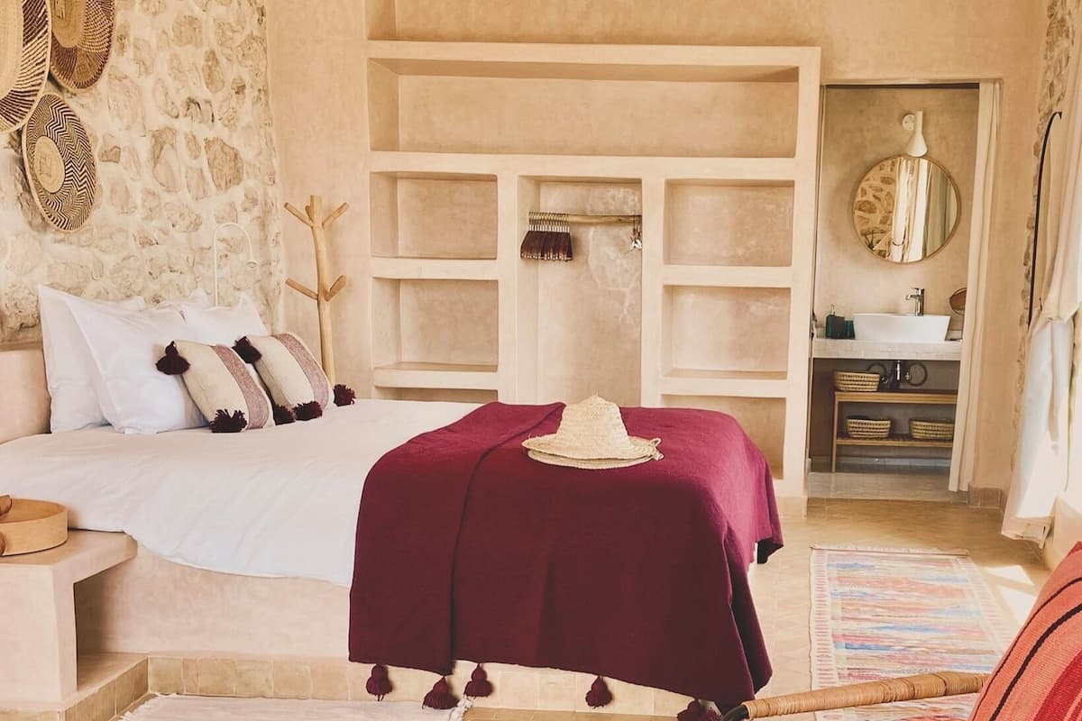 A bedroom with a bed, a bedside table, a chair, a small table with a vase of flowers, and a bathroom visible through an open door.