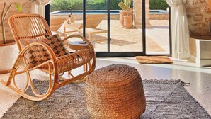 Una stanza con una sedia in vimini, un poggiapiedi intrecciato e vista su una piscina e vegetazione attraverso una porta vetrata.