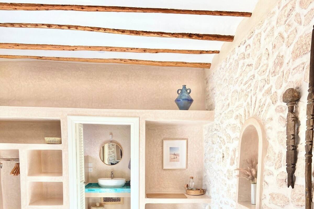 A cozy bedroom with a stone wall, a wooden ceiling, a bed with patterned bedding, a bedside table, and a small wooden stool.