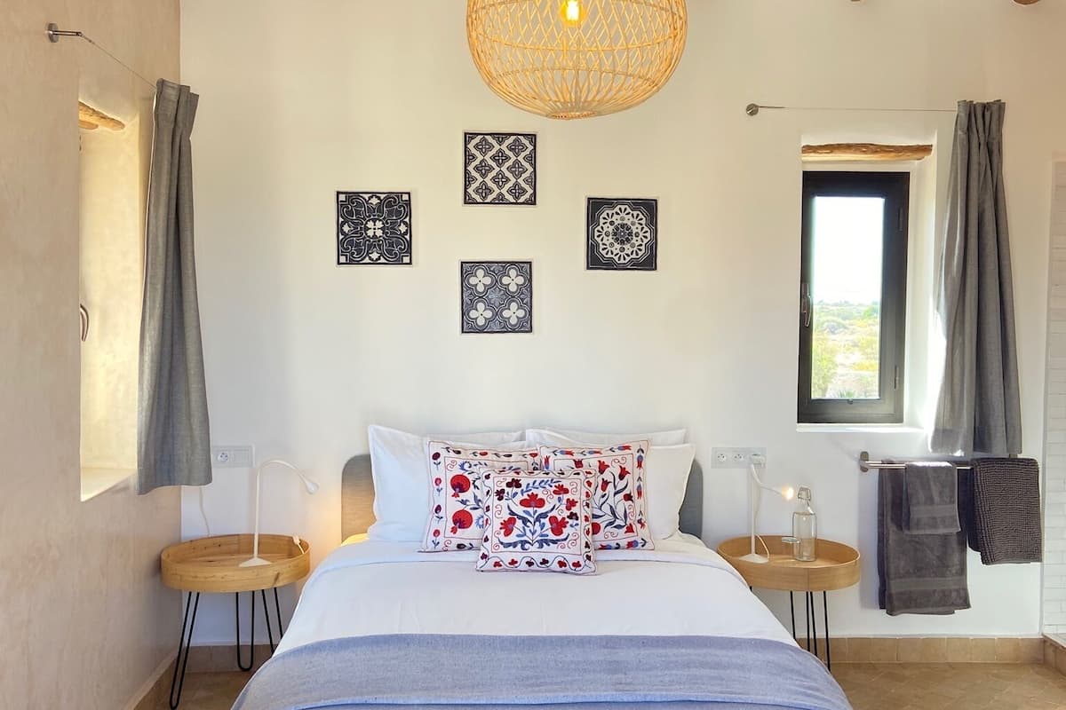 A bedroom with a bed, a hanging light fixture, a window, and a wall with framed artwork.