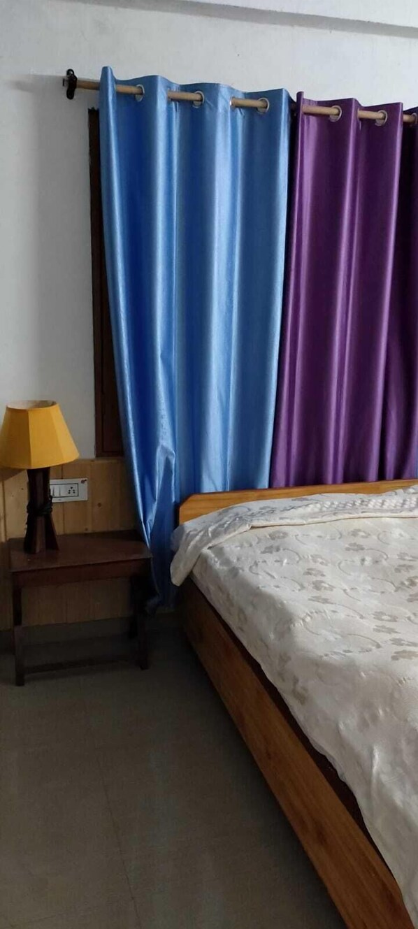 A bedroom with a bed, a nightstand with a lamp, and a window with blue and purple curtains.