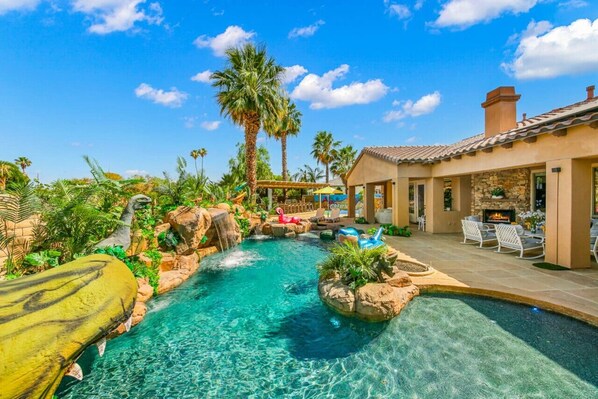 A pool area with a waterfall, lounge chairs, and a stone building with a fire pit.