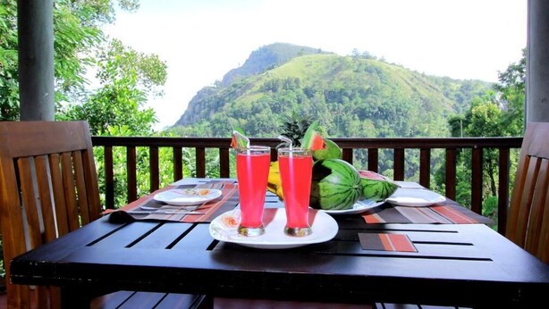 A table with two drinks, a watermelon, and a plate of food on a balcony with a mountain view. - Blue Ribbon Homestay  (Ella)