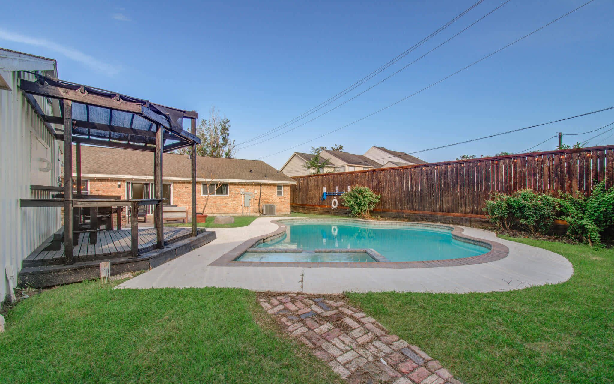 A backyard with a swimming pool, a wooden deck, and a brick patio.