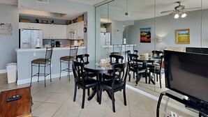 A modern kitchen with white cabinets, a refrigerator, and a coffee maker. There is a dining area with black chairs and a glass table. A TV is mounted on the wall.