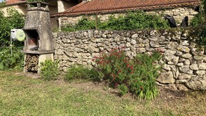 A stone wall with a rustic stone oven, lush greenery, and a building with a tiled roof.