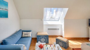 Une chambre à coucher confortable aménagée en mezzanine, avec un plafond en pente, une grande lucarne, un canapé bleu, une petite table sur laquelle se trouve une théière rouge, et des boîtes de rangement décorées sur le thème de la marine.