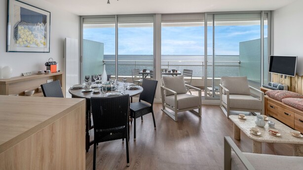 Una habitación moderna con zona de comedor, sofá y televisión. Cuenta con ventanas amplias con vista al mar.